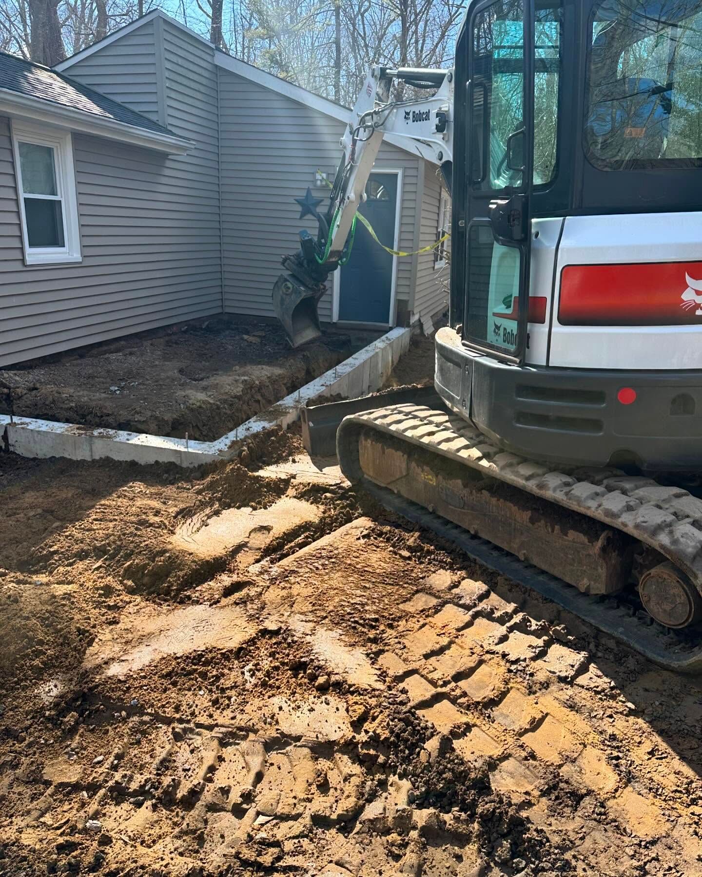 A small excavator digs a trench next to a house with a blue door.