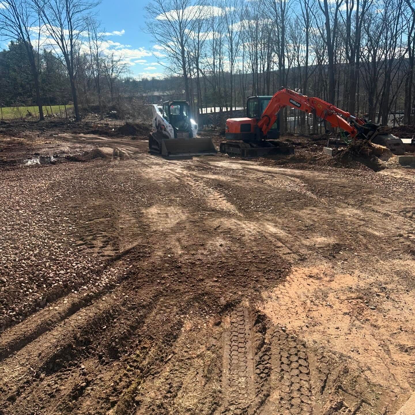 Construction site with a Bobcat and an excavator working on dirt. Trees and blue sky in background.
