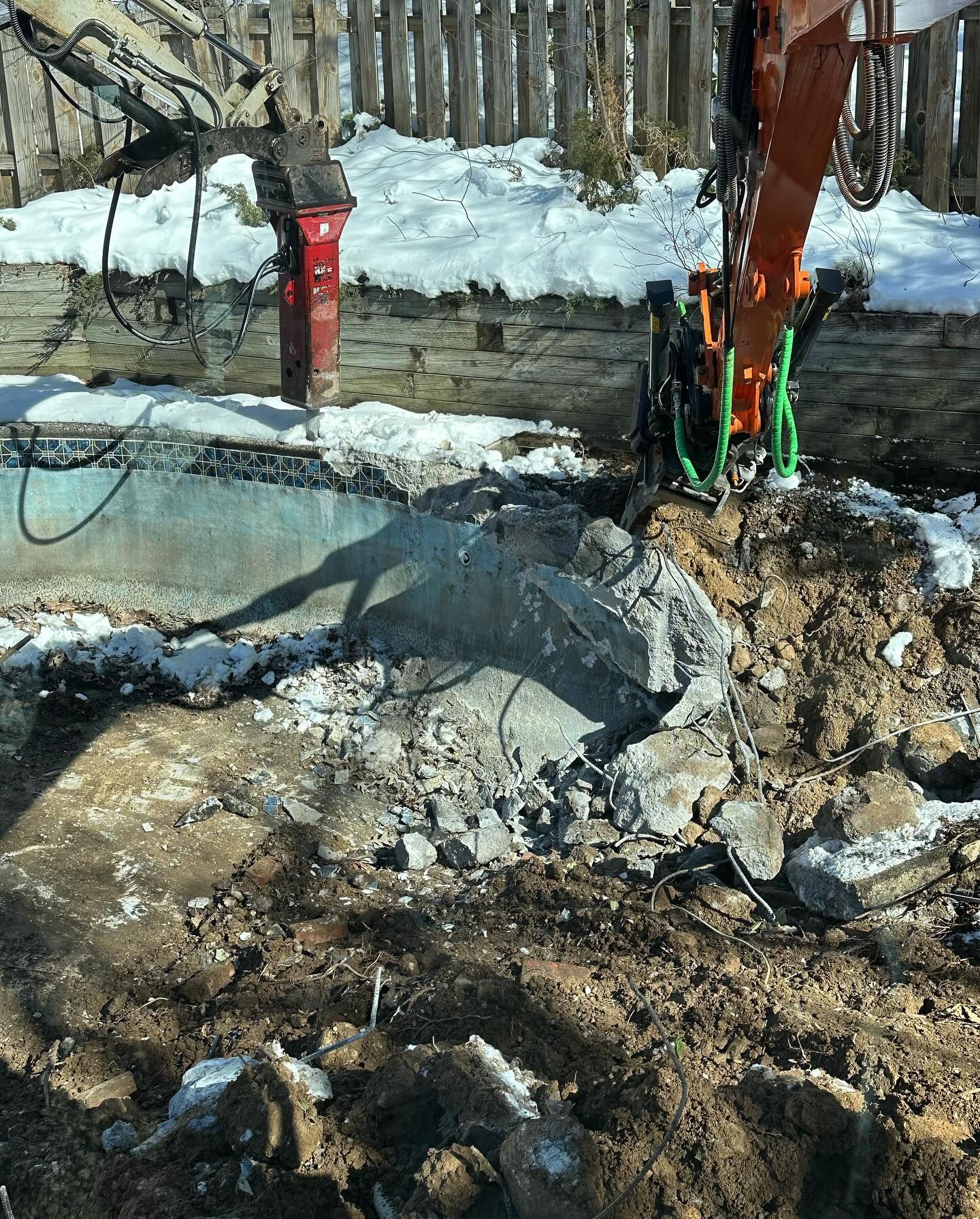 An excavator demolishes a concrete structure outdoors with snow on the ground, sunshine and a wooden fence in background.
