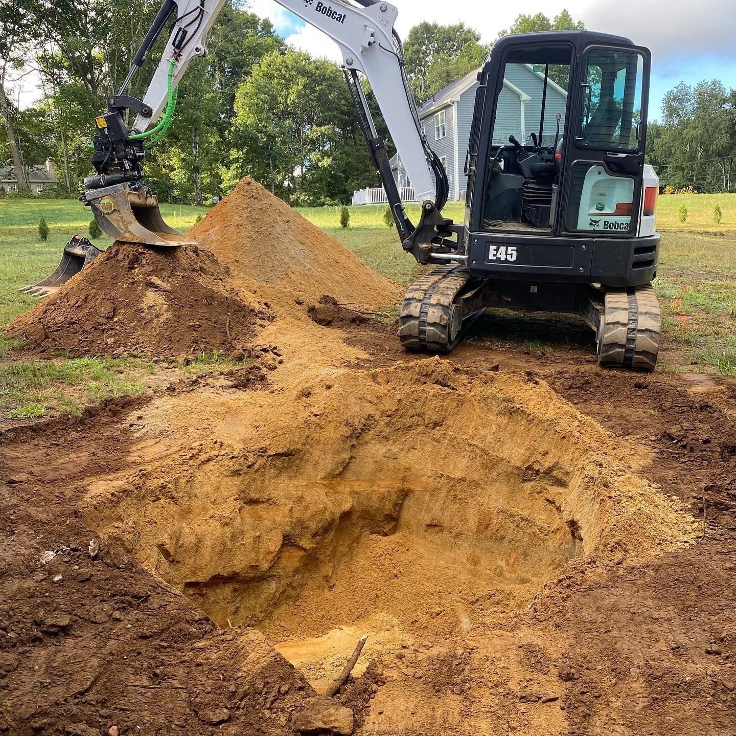 Mini-excavator digging a large hole in a grassy yard, with a pile of dirt beside it.