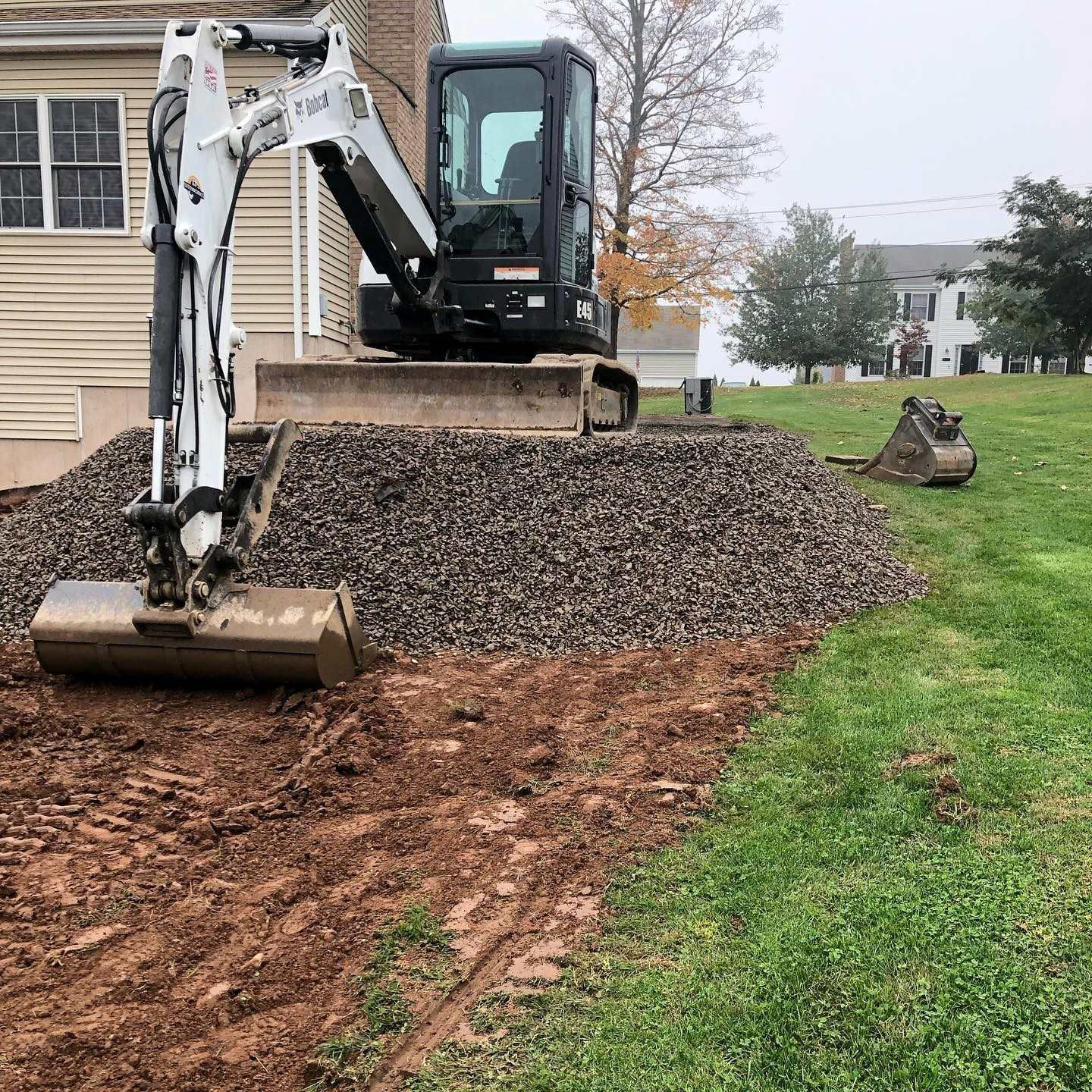 Mini excavator on gravel pile, digging earth next to a building on a grassy area.