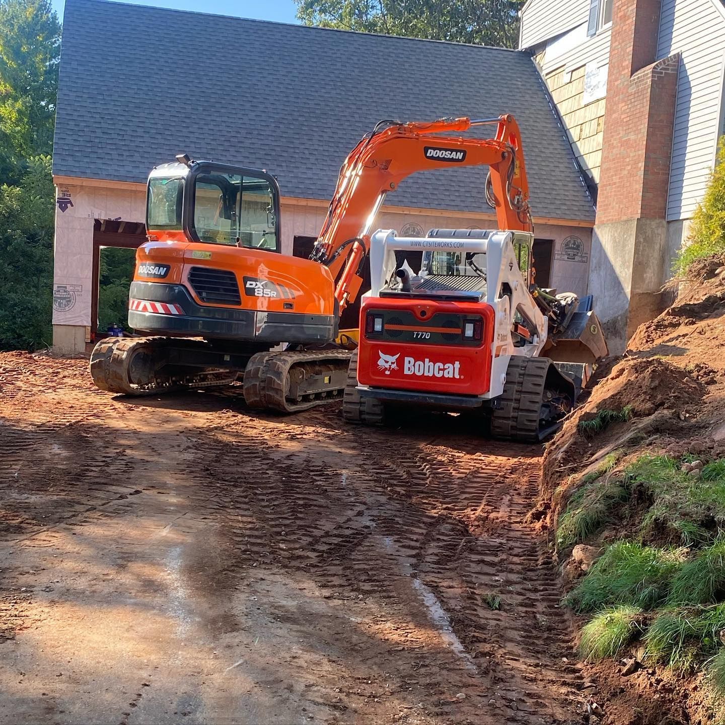 Orange excavator and Bobcat skid-steer on a muddy construction site near a building.