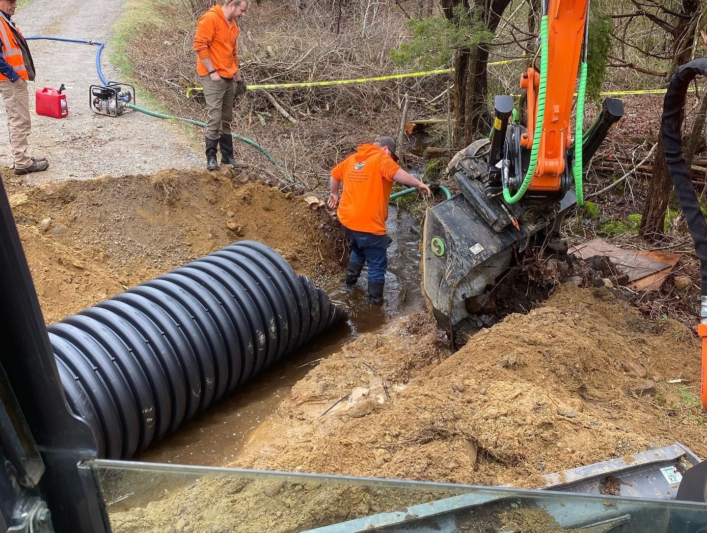 Workers installing a large corrugated pipe. An excavator and two people are visible in a muddy ditch, orange safety shirts.