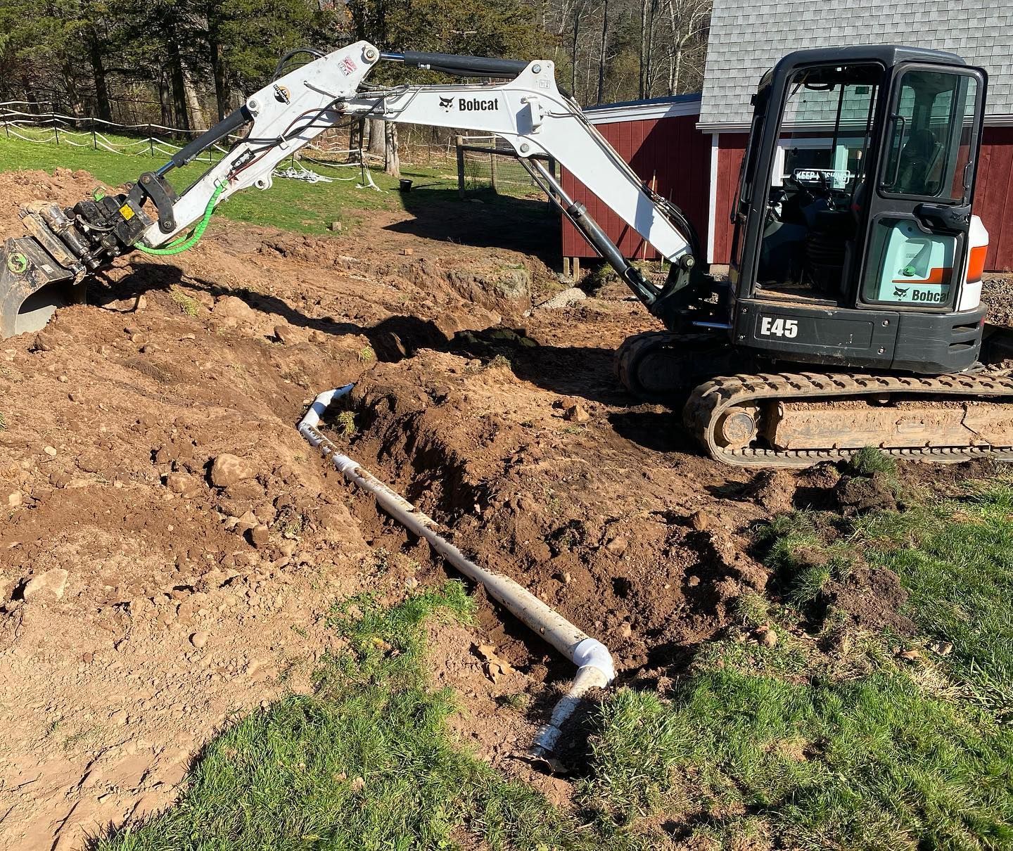 Mini excavator digging a trench around a white pipe on a grassy area near a red building.