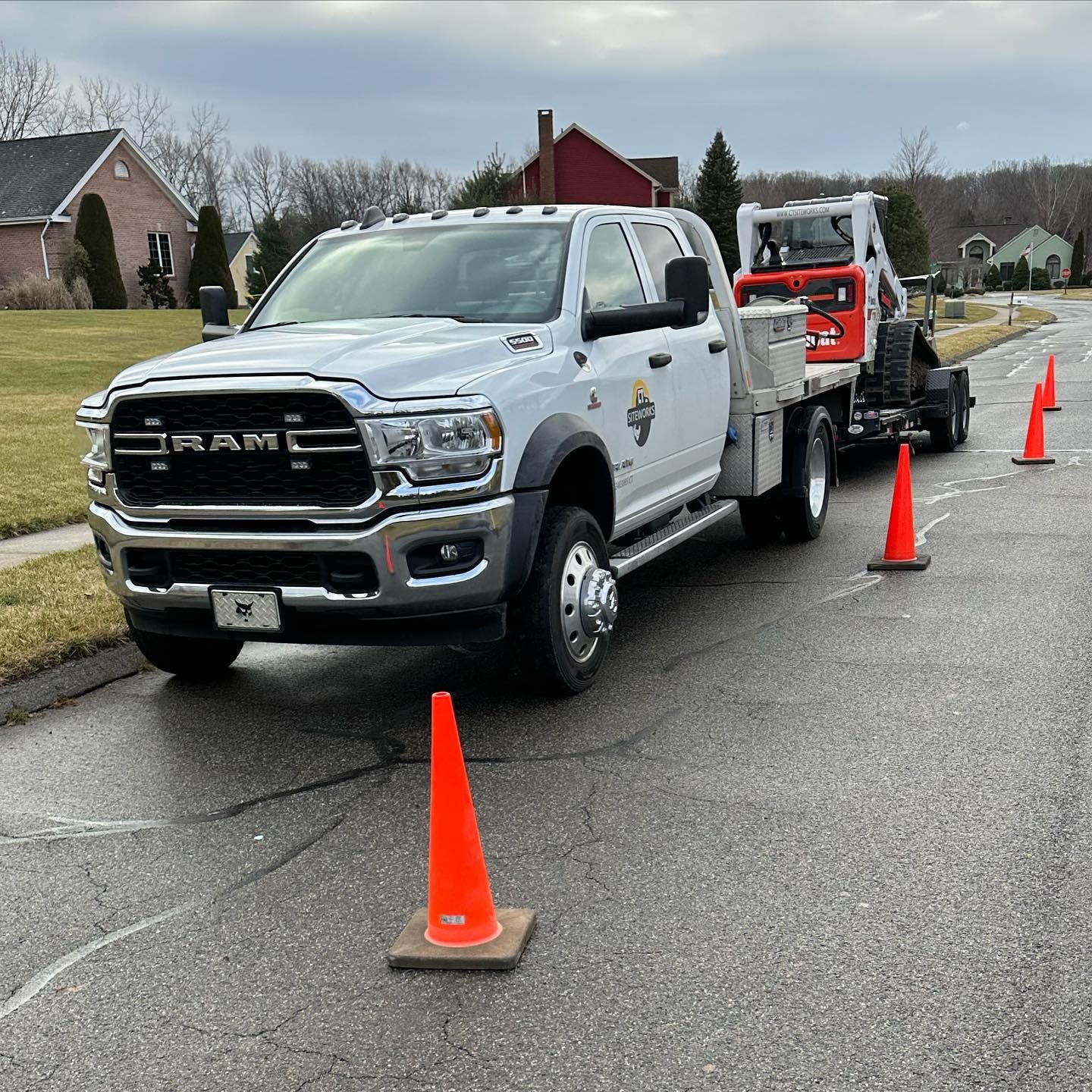 White RAM truck with trailer carrying a skid steer on a residential street; orange cones mark the roadside.