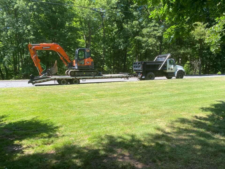Dump truck towing an excavator on a trailer parked on a grassy area, trees in the background.