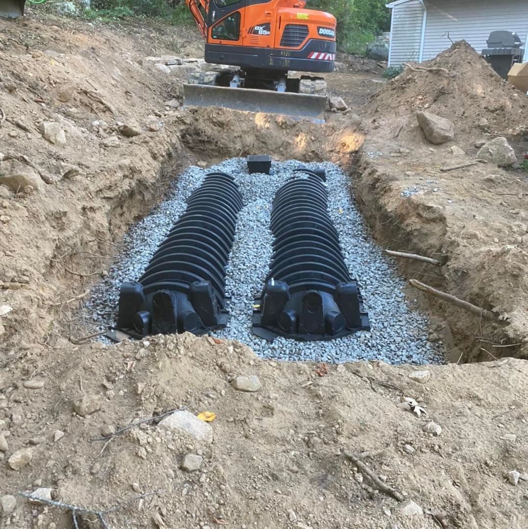Trench with two black septic tanks surrounded by gravel and soil, construction equipment in background.