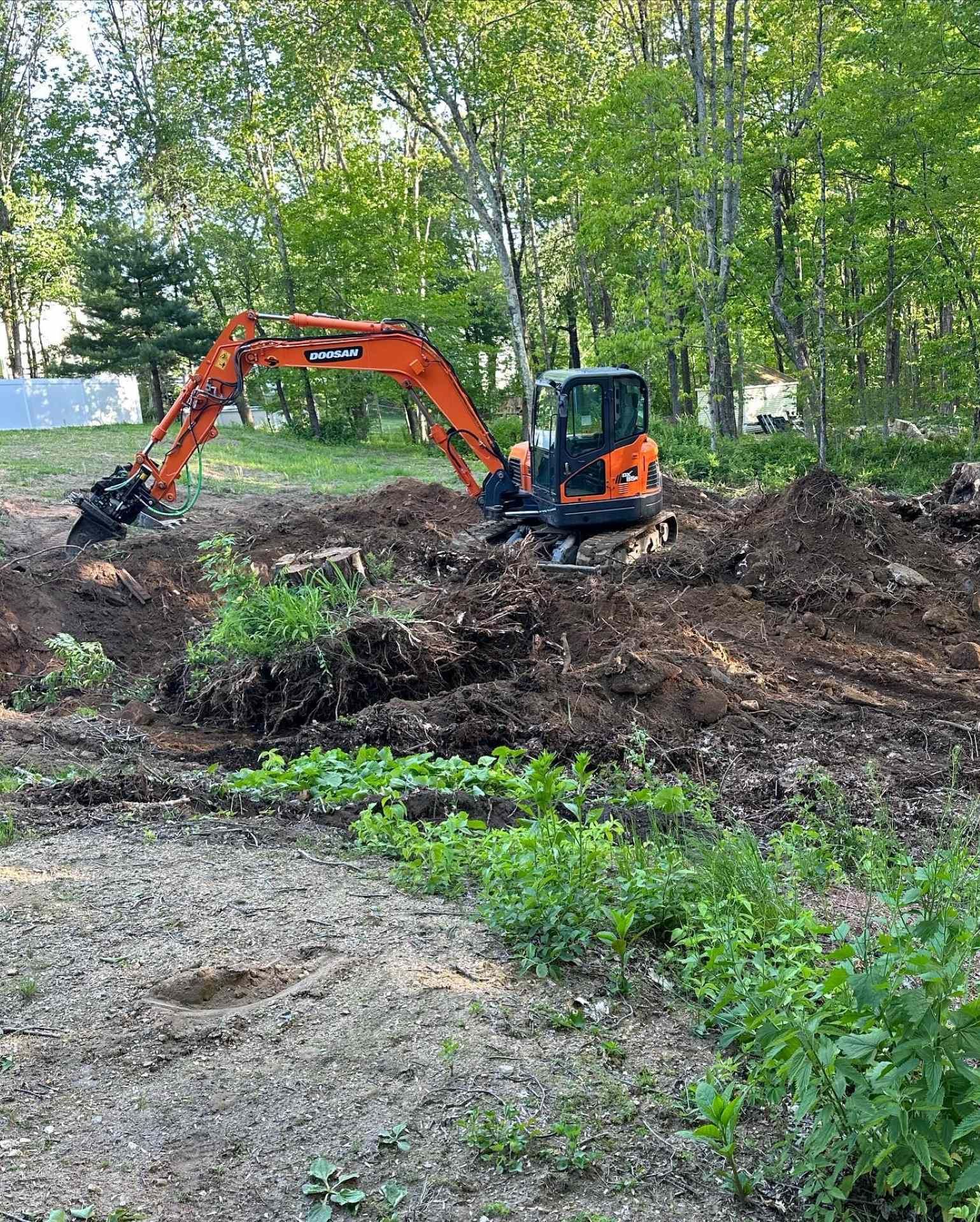 An orange excavator digging in a dirt area surrounded by green plants and trees.