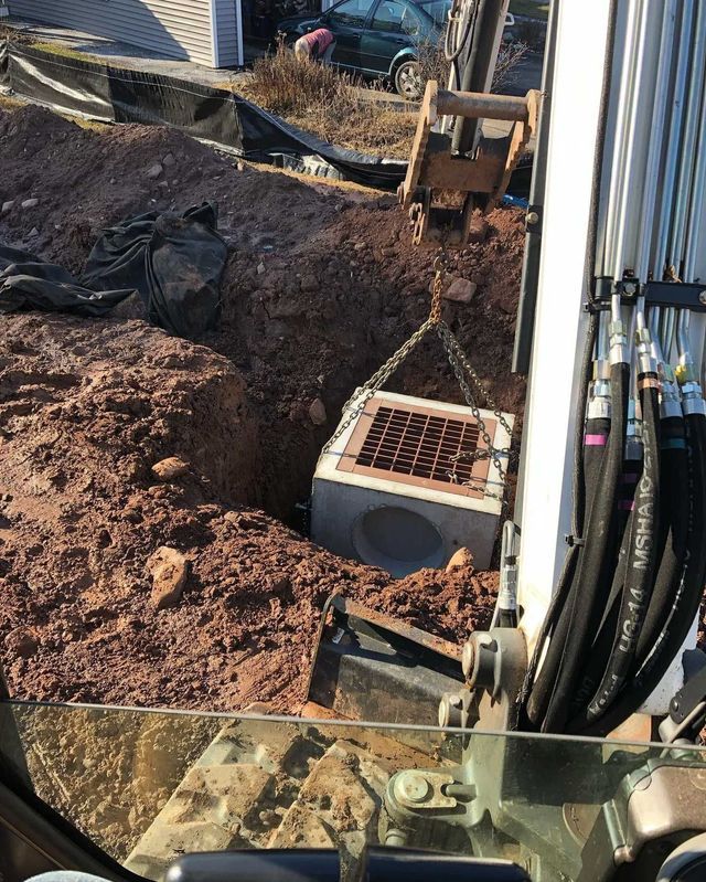 An excavator lowering a concrete drainage culvert into a red dirt pit.