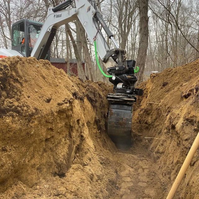 Bobcat excavator digging a trench in a wooded area, with dirt and trees visible.
