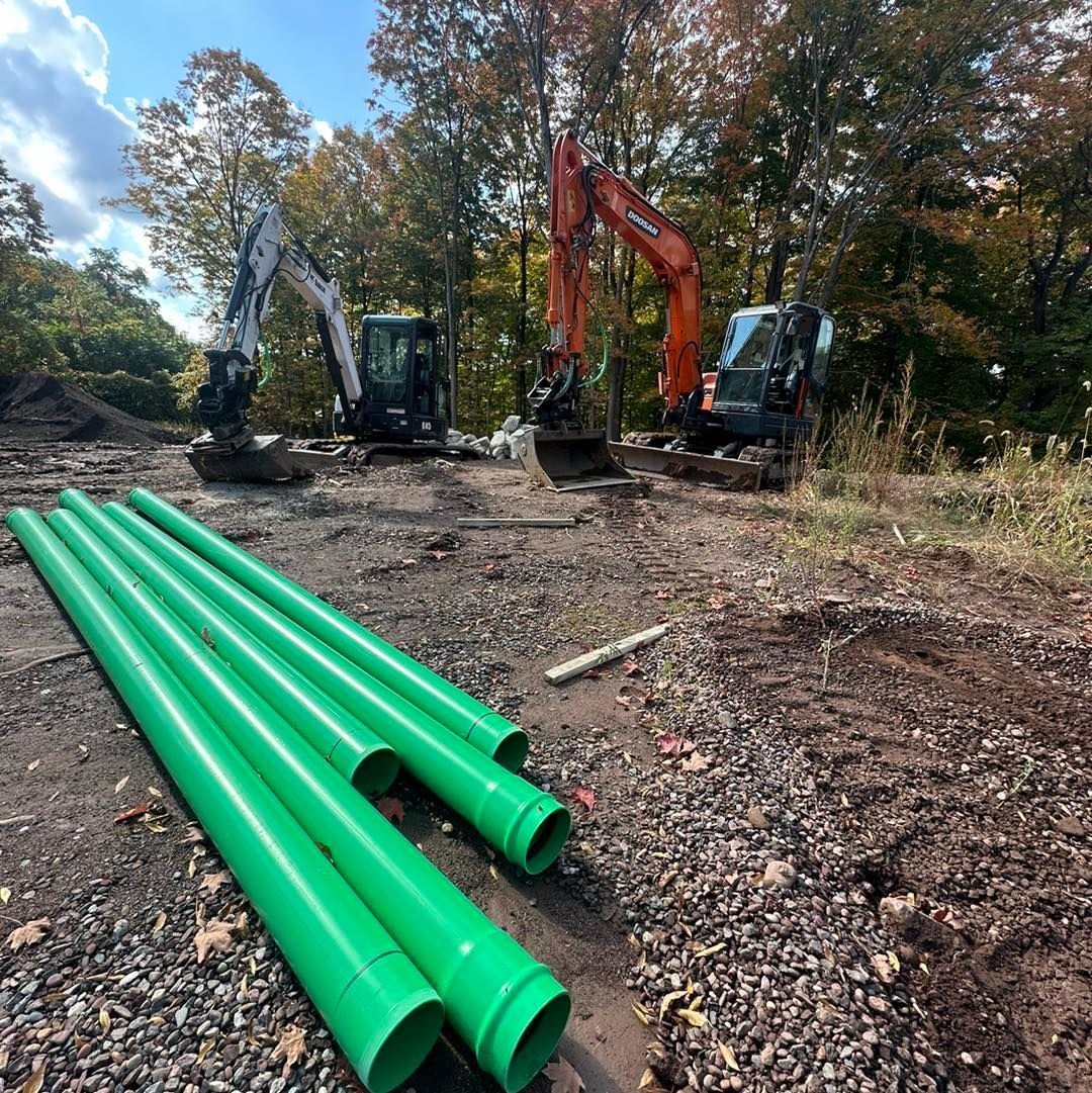 Green pipes on ground, excavators digging in construction site, forest backdrop.