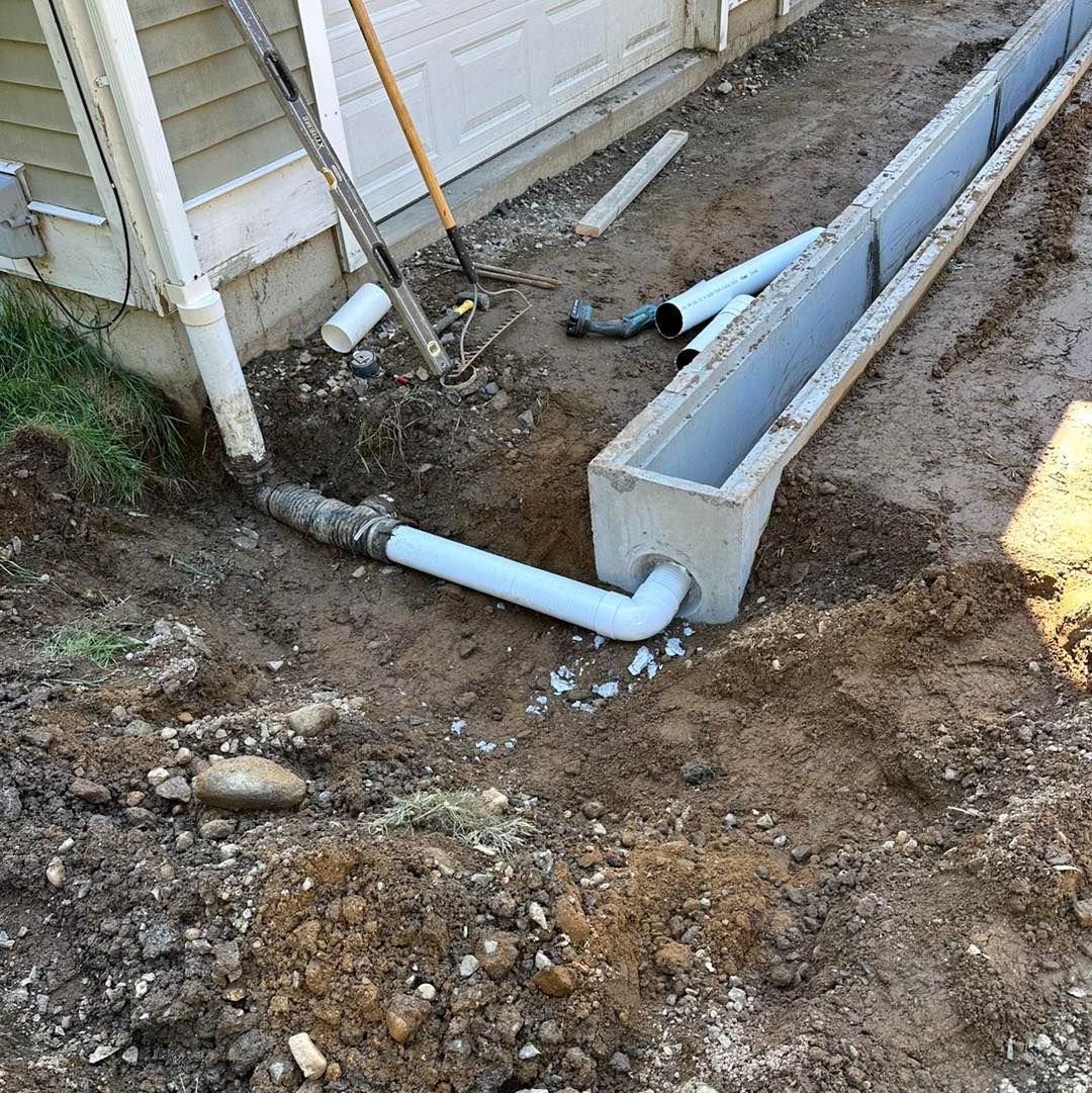 Construction site: concrete trench drain connected to a downspout with PVC pipe, partially buried.