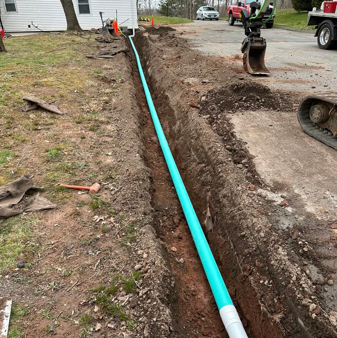 A turquoise pipe in a trench runs along a driveway. Construction equipment is nearby.