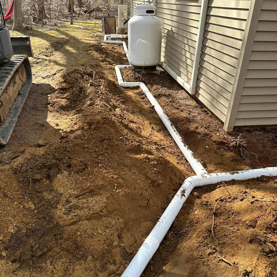 Trench with white pipes leading from a propane tank to a house exterior. A small excavator is in the frame.