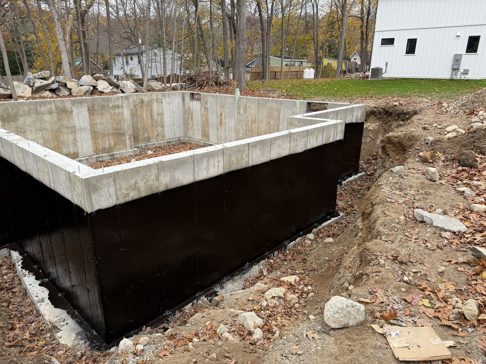 Concrete foundation with black waterproofing coating, surrounded by dirt and leaves, outdoors.