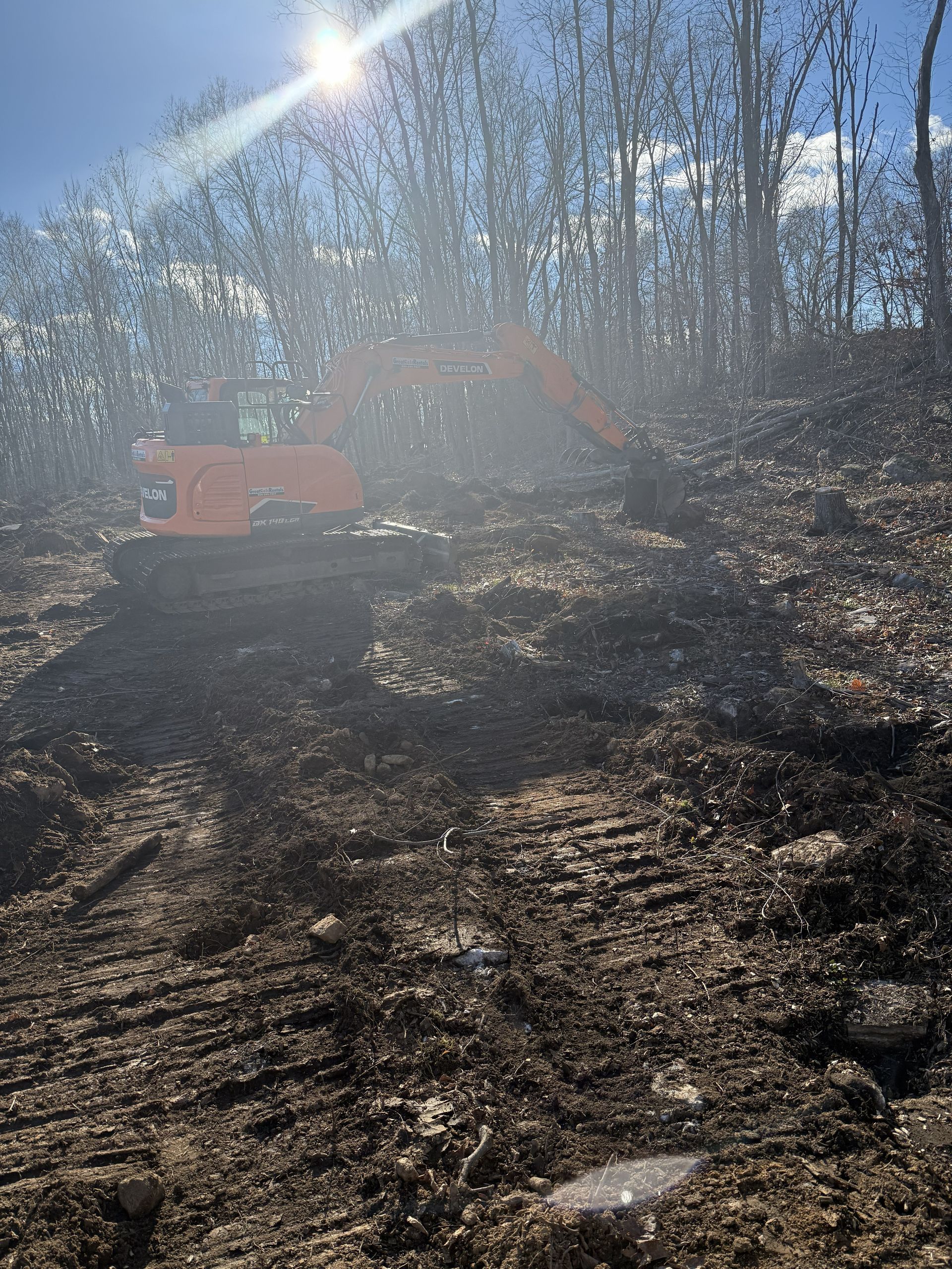 An orange excavator on a dirt hill clearing land. Trees and sun in background.