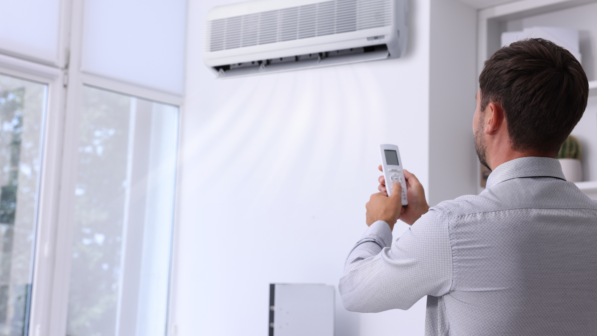 Man using remote control to operate air conditioner, white wall, window.