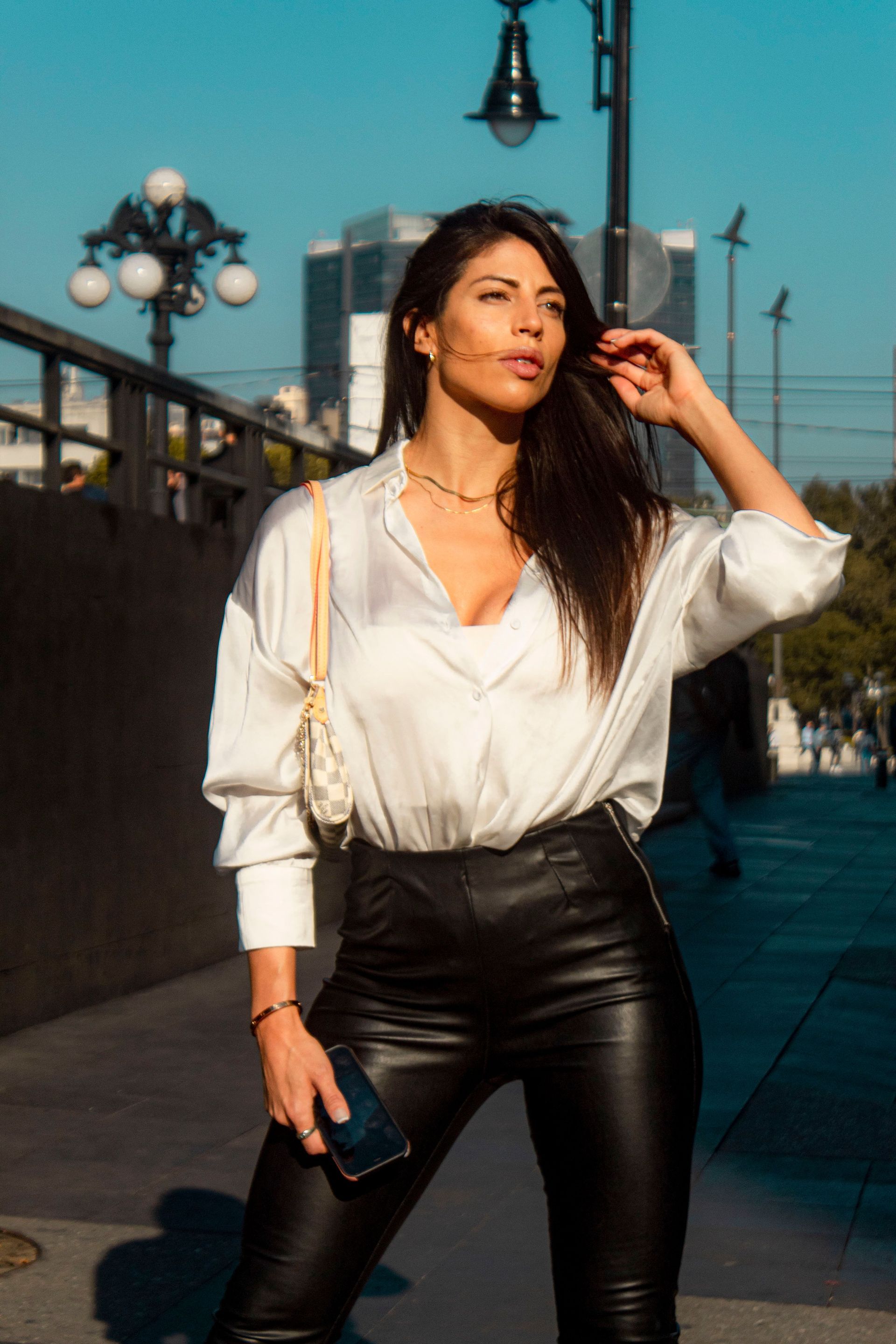Mujer con blusa blanca y pantalones negros posando al aire libre en una calle soleada.