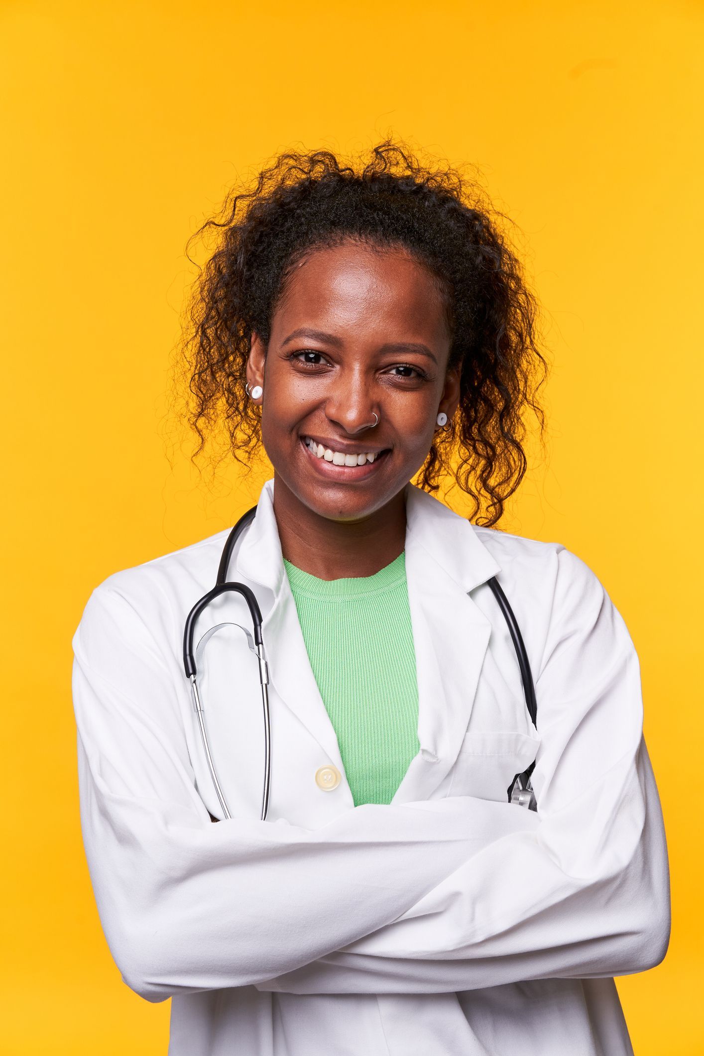 A female doctor with a stethoscope around her neck is smiling with her arms crossed.