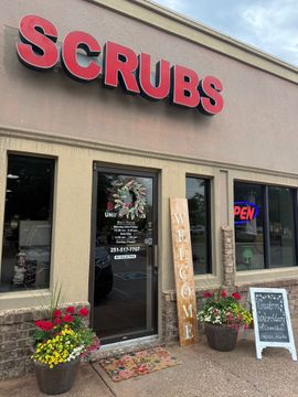 Scrubs storefront with red sign, glass door, flower planters, and an open neon “OPEN” sign.