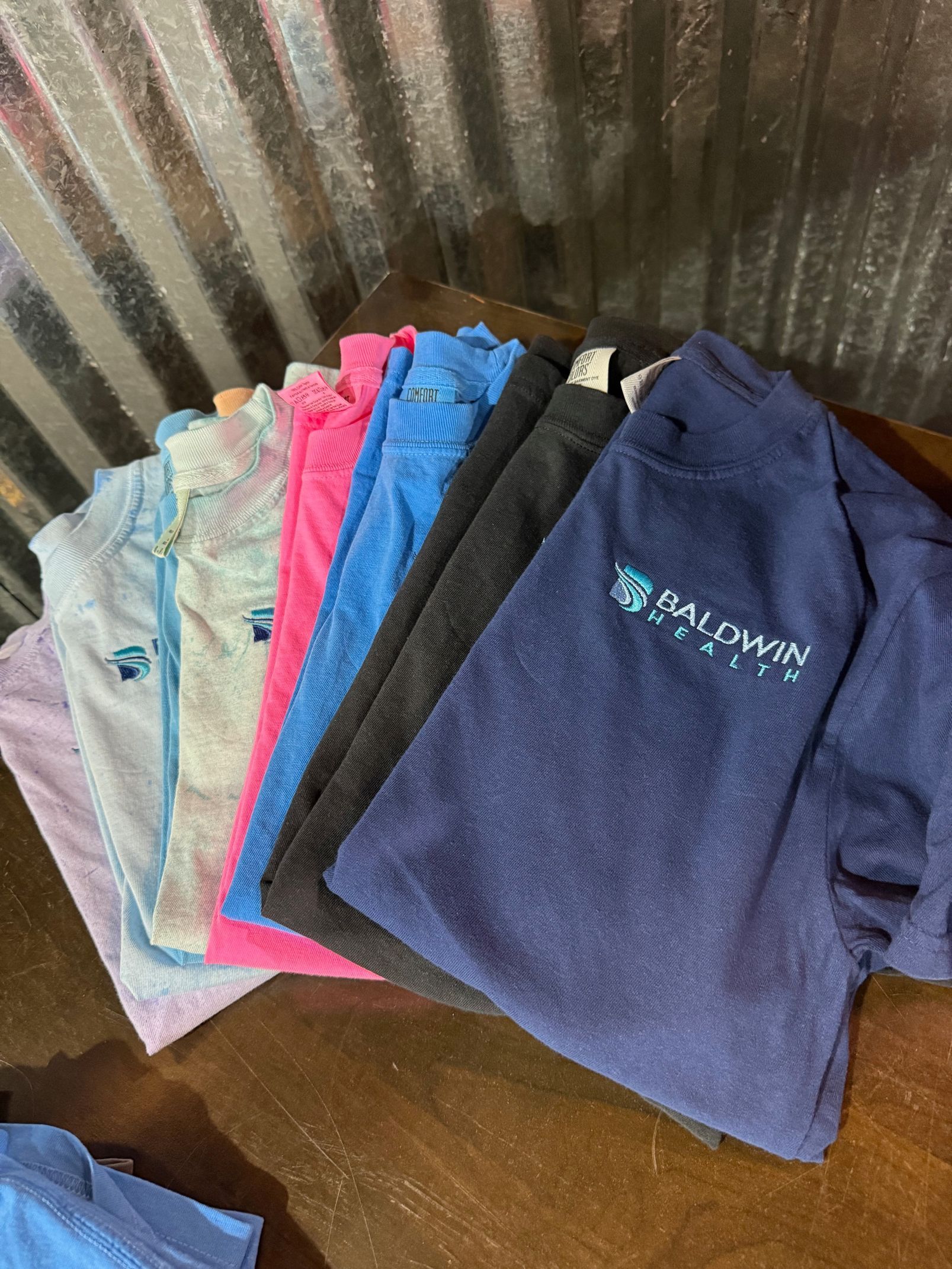 Stack of folded T-shirts in various colors, including navy, gray, pink, and blue, on a brown surface.