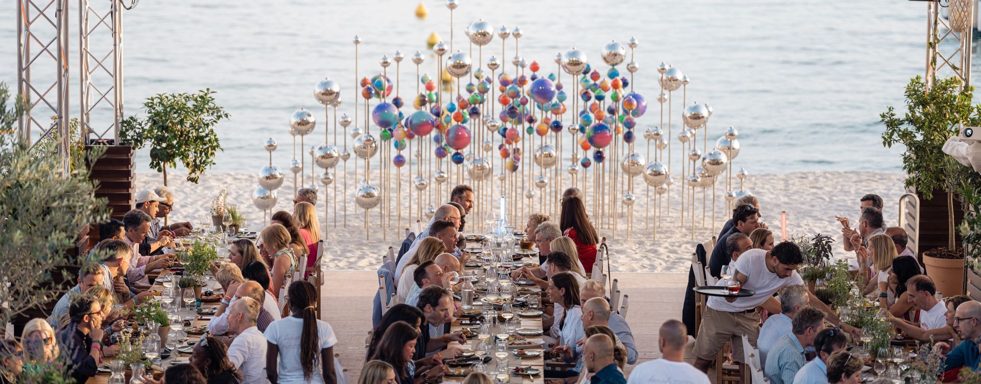 A large group of people are sitting at a long table on the beach.