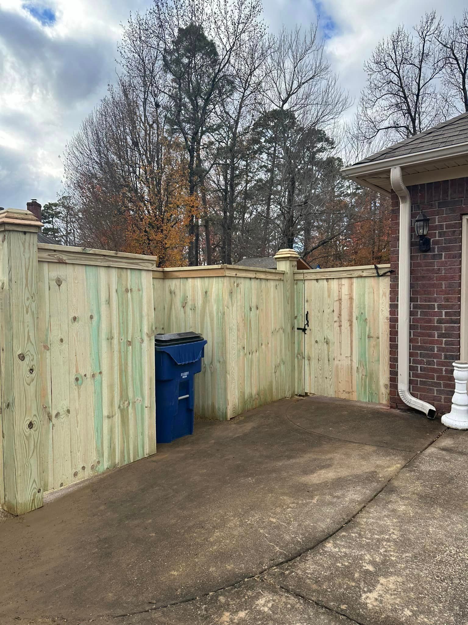 A wooden fence with a blue trash can in front of a brick house.