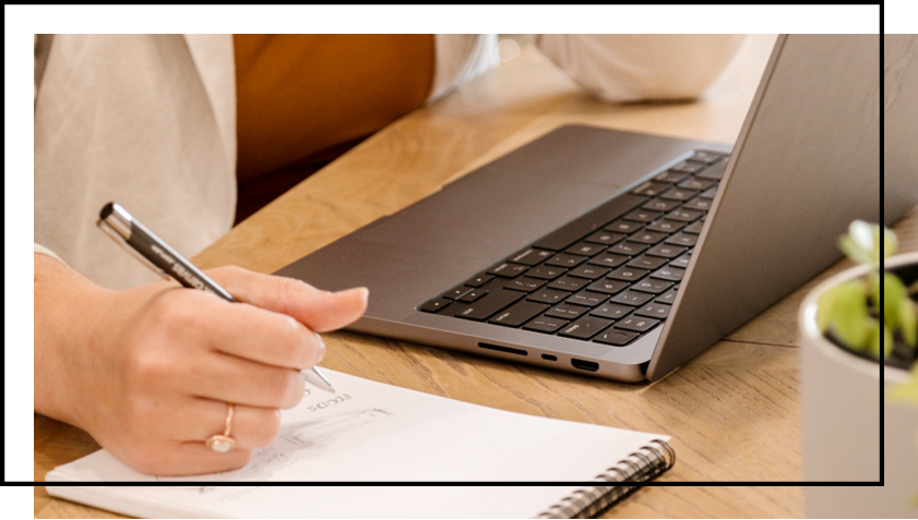 A person is writing in a notebook in front of a laptop computer.