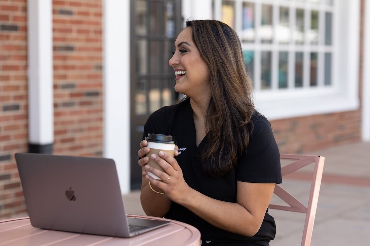 Woman with coffee laughs while looking away from a laptop at an outdoor table.