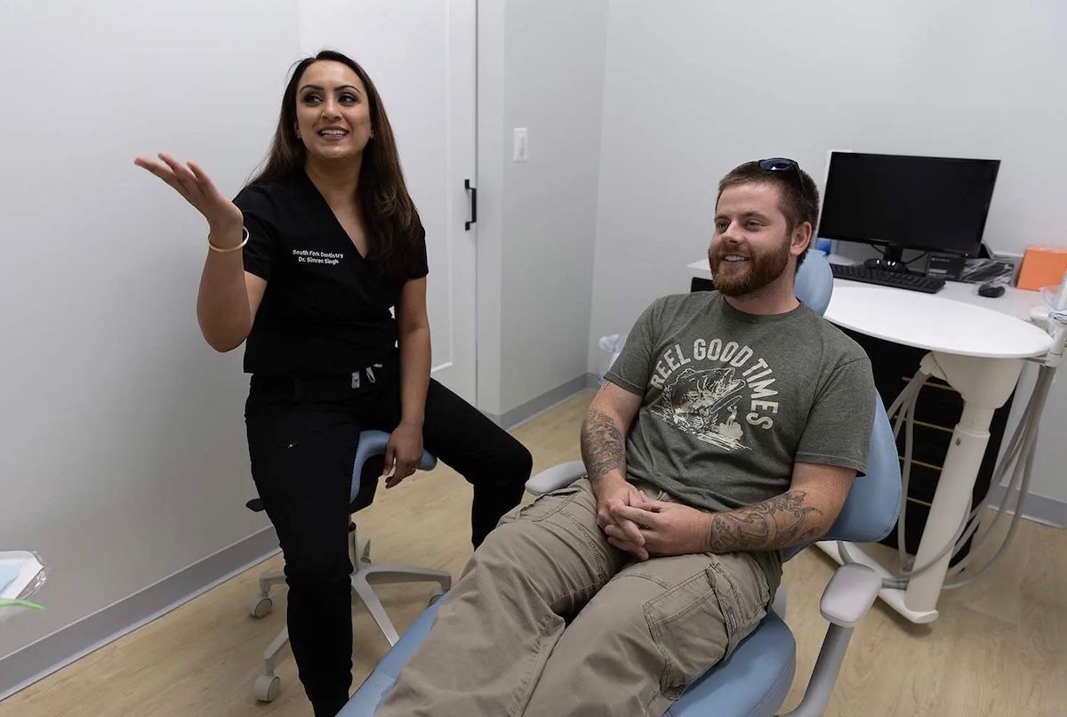 Dentist in black scrubs explaining something to a smiling male patient in dental chair.