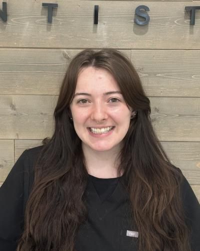 Woman with long brown hair smiling, wearing black scrubs in front of a wooden wall.