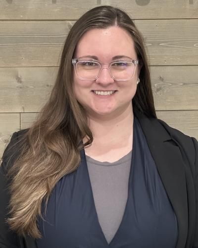 Woman with long brown hair, glasses, and a blazer smiling in front of a wooden wall.