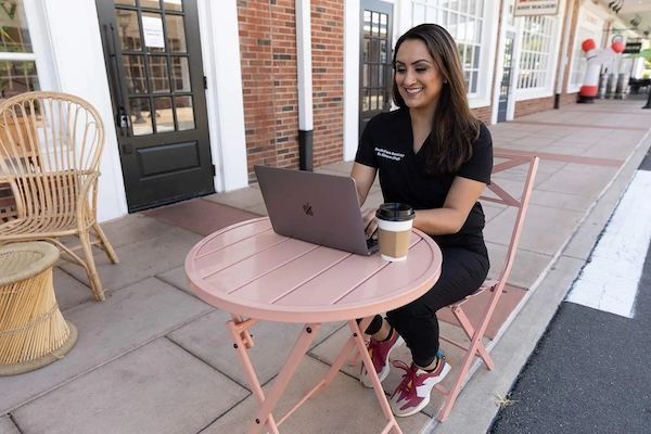Woman in black scrubs smiles, works on laptop at pink outdoor table, holding coffee.