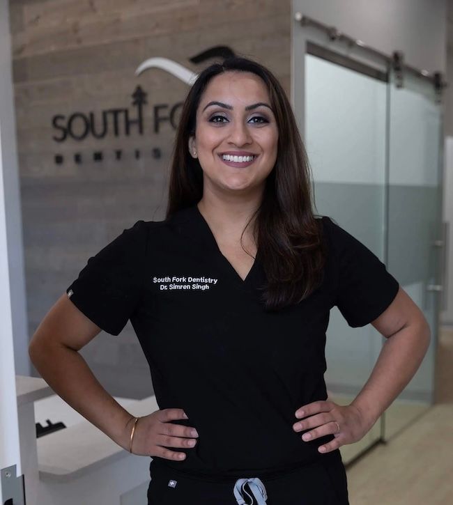 Woman in black scrubs with arms akimbo smiles in a dental office.
