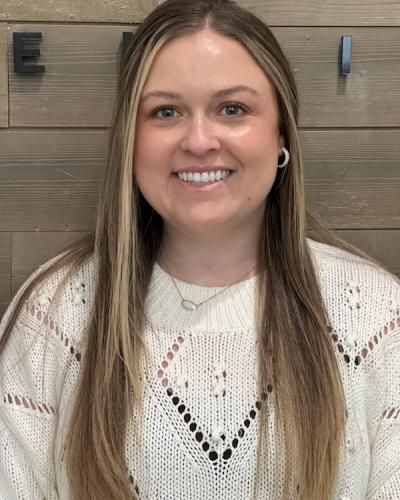 Woman with blonde hair smiling, wearing a cream sweater and necklace, in front of a wood-paneled wall.