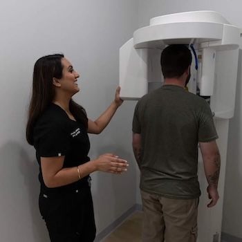 A dentist assists a patient with a dental X-ray machine. The setting is a dental office.
