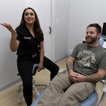 Dentist in black scrubs consults with a male patient in khaki pants and a green shirt in a dental office.