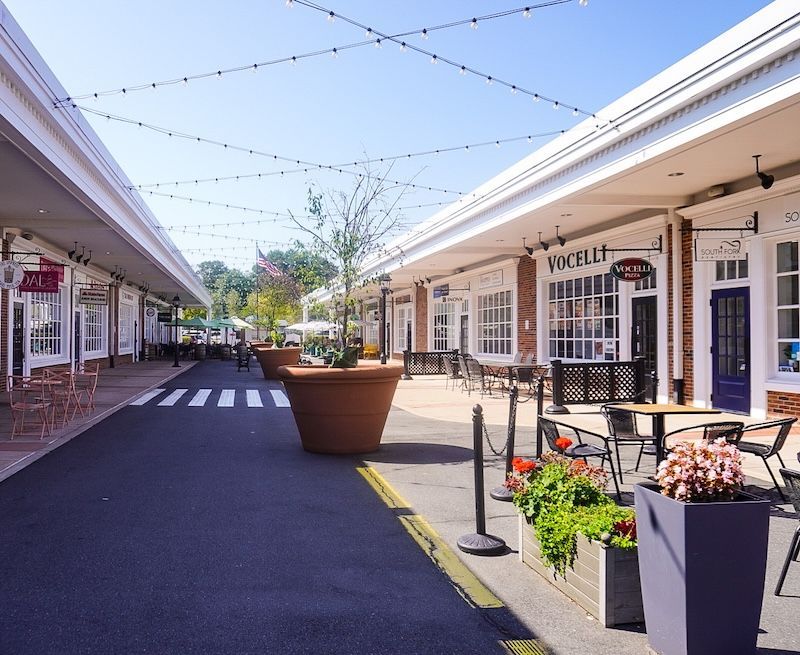 Outdoor shopping area with brick buildings, string lights, and seating. Sunny day.