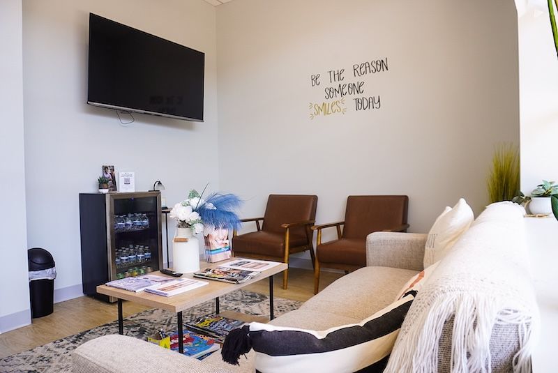 Waiting room with sofa, chairs, TV, drinks, magazine, and a quote on wall.