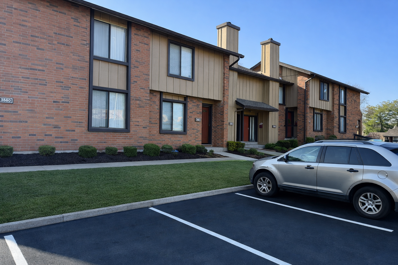 Apartment building exterior with a parked SUV in front. Brick and tan siding, blue sky.