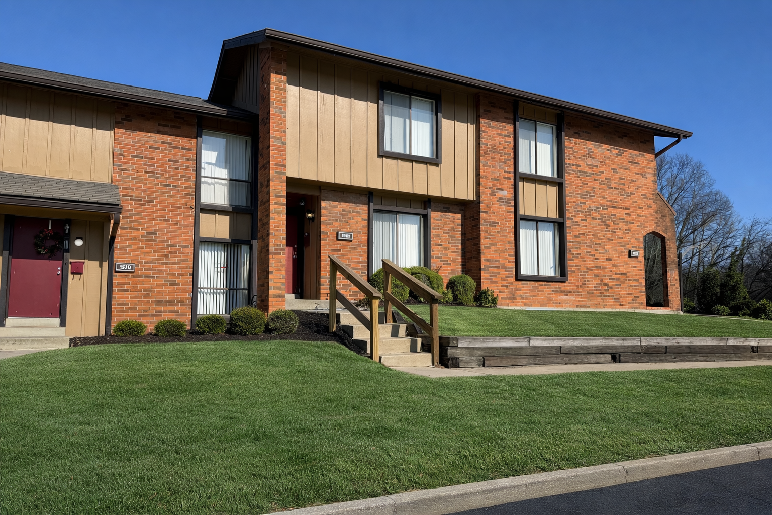Two-story brick townhomes with brown trim and roof, set on a green lawn, and a clear blue sky.