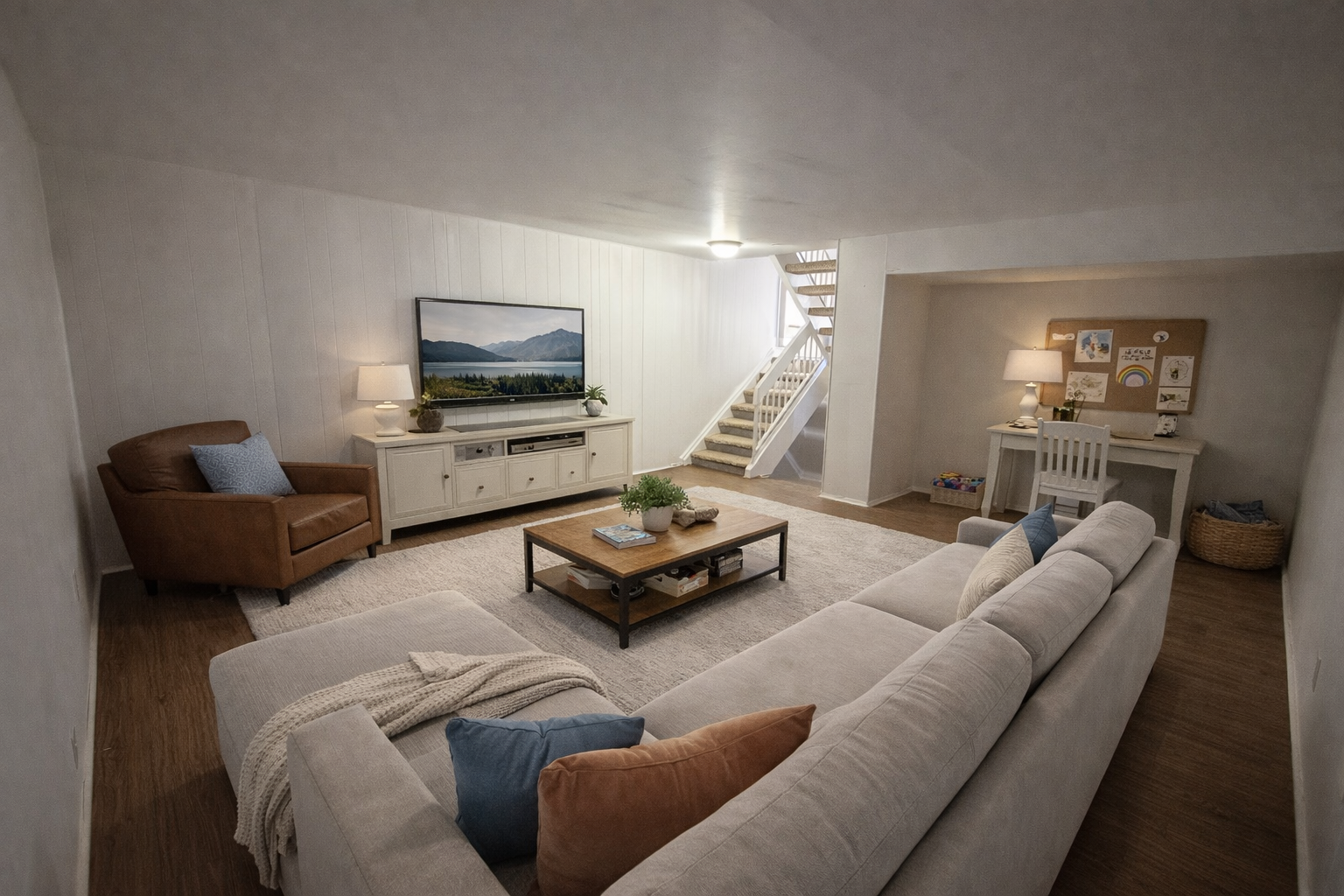 Cozy basement living room with a sectional sofa, TV, desk, and staircase. Neutral colors.