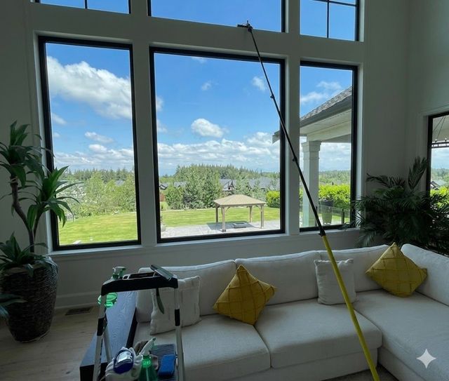 Bright living room with large windows overlooking a green field and blue sky, white sofa, and potted plants.
