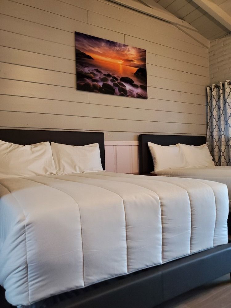 Two beds with white bedding in a room with a sunset picture on the wall.