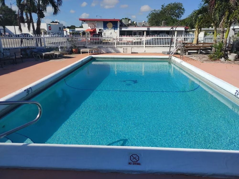 Pool with blue water surrounded by a red deck and white fence, in front of a building under a sunny sky.
