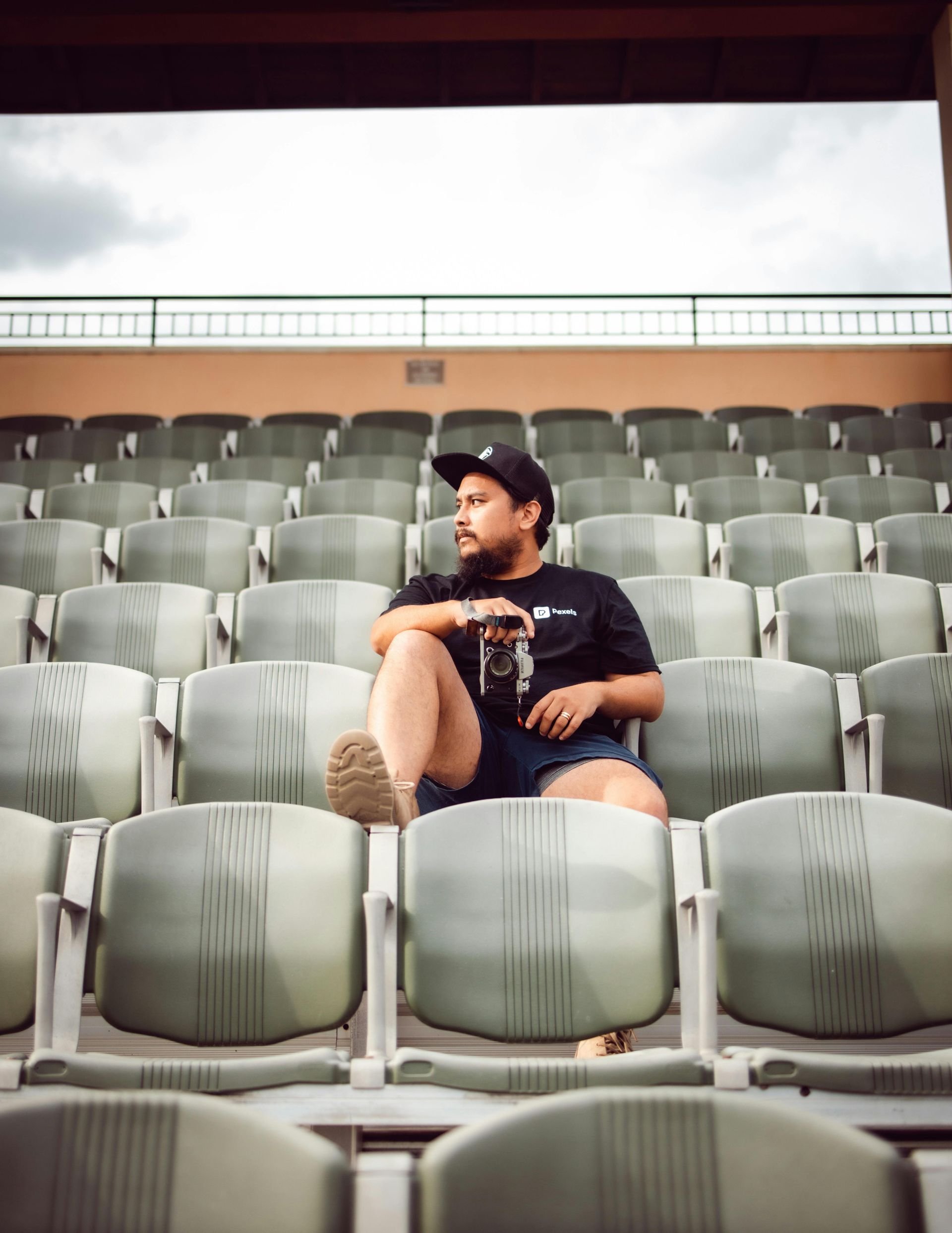 Man in baseball stadium seat, looking away, holding a camera. Wearing a black shirt, shorts, and hat.