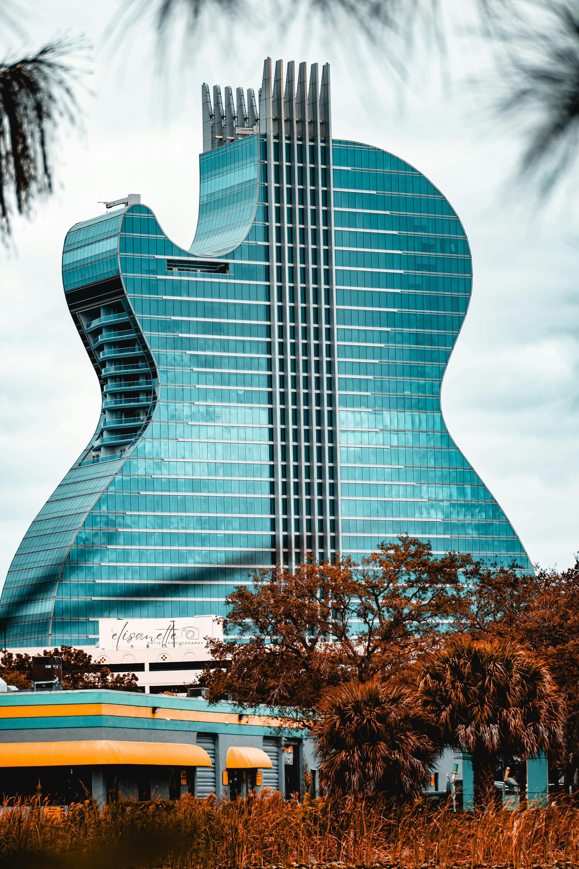 Guitar-shaped Hard Rock Hotel building with teal glass exterior, seen amidst trees and a cloudy sky.