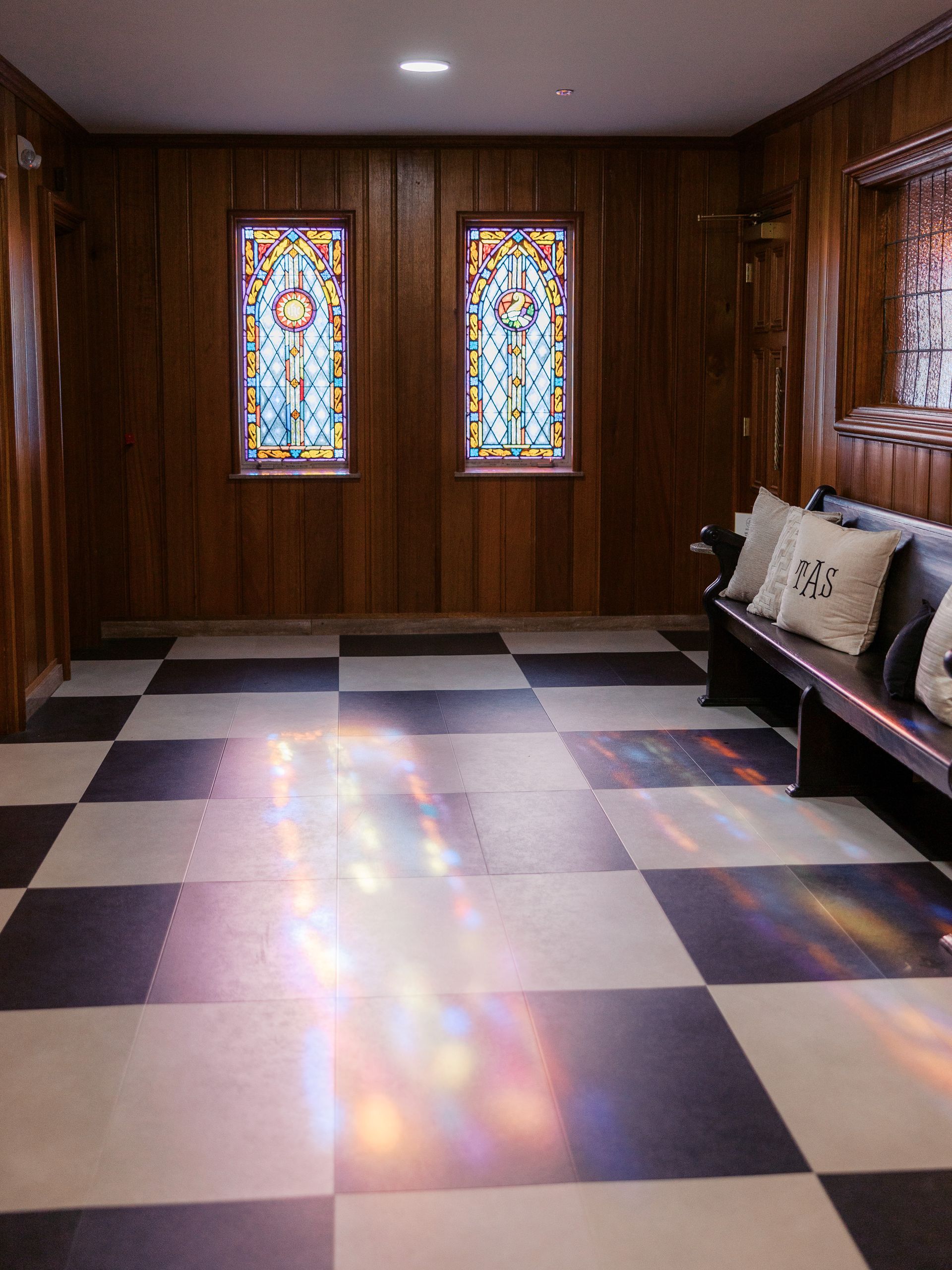 Hallway with stained glass windows, checkered floor, wood paneling, and a bench with pillows.