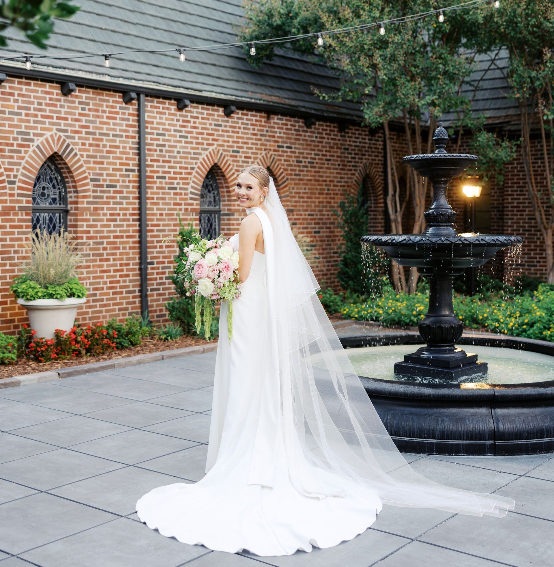 Bride in white dress, holding bouquet, poses by a fountain in front of a brick building.
