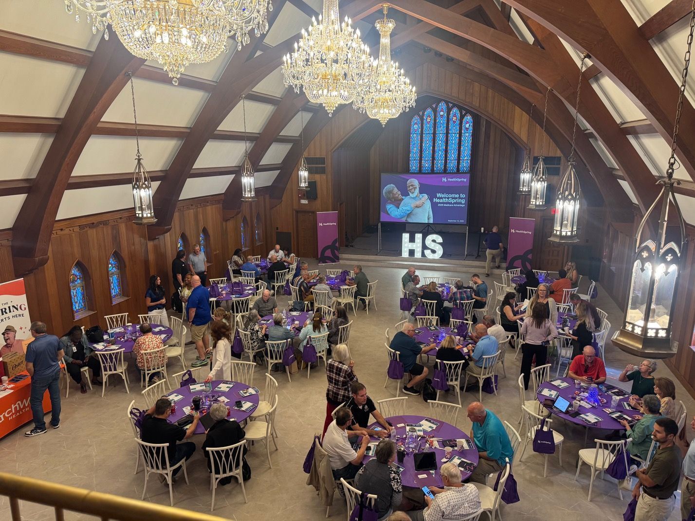 Event in a large, high-ceilinged room with tables, attendees, and a presentation screen. Purple accents and logos.